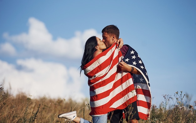 Beijando um ao outro. Sente liberdade. Lindo casal com bandeira americana se diverte ao ar livre no campo.