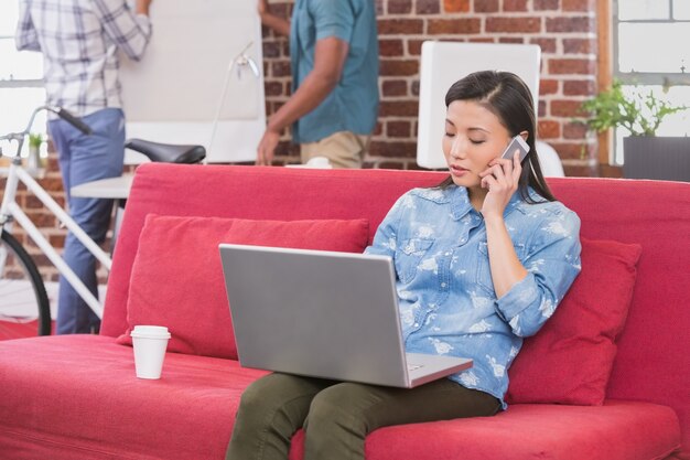 Beiläufige Frau, die Laptop und Handy auf Couch verwendet