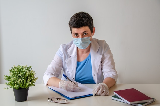 Beim Arzttermin. Ernsthafte Therapeutin in Schutzmaske schreibt Diagnose auf und sitzt am Schreibtisch gegenüber dem Patienten im Büro