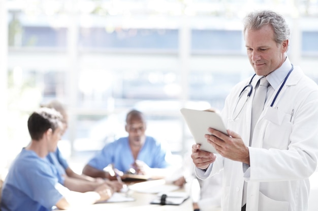 Beim täglichen Check Ein leitender Arzt hält ein Touchpad, im Hintergrund sitzen Kollegen