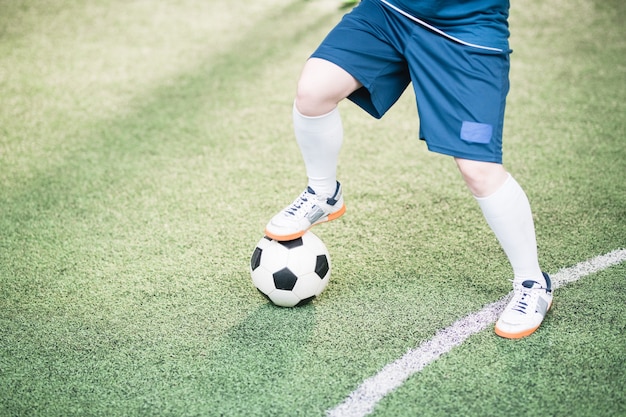 Beine der jungen aktiven Spielerin in der blauen Uniform, die den rechten Fuß auf Fußball während des Fußballspiels auf dem Feld hält