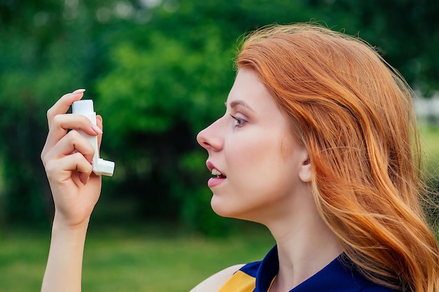 Bekämpfung von Asthma und Bronchitis. Aktive, fröhliche, schöne, junge Ingwer-Rothaarige, irische Norwegerin in einem gelben Kleid, die den Inhalator im Sommerpark verwendet. Konzept der Frühlingsallergie.