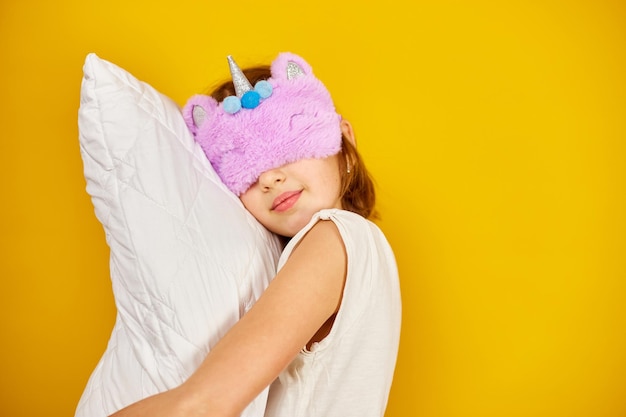 Foto bela adolescente de pijama branco com uma máscara de dormir violeta abraça um travesseiro