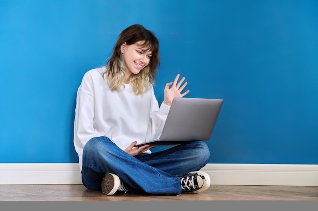 Bela adolescente sentada no chão com o laptop sobre fundo azul