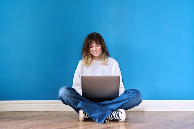 Bela adolescente sentada no chão com o laptop sobre fundo azul