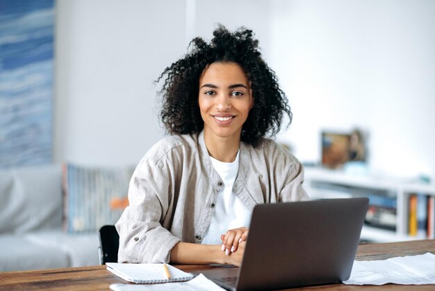 Foto bela, amigável, confiante, jovem mulher afro-americana em roupas casuais elegantes, gerente freelancer ou estudante sentada em um laptop, trabalhando ou estudando em casa, olhando para a câmera sorrindo.