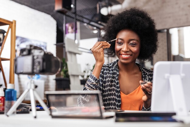 Bela amostra. mulher sorridente de pele escura em um top laranja sorrindo positivamente enquanto experimenta o novo formato de sobrancelha