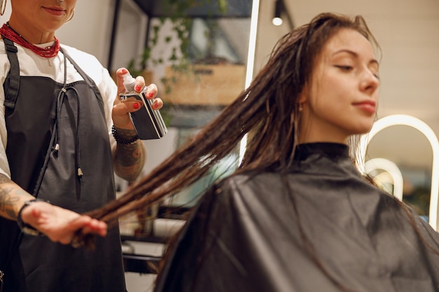 Bela cabeleireira com mãos tatuadas aplicando spray no cabelo molhado da mulher no salão de beleza