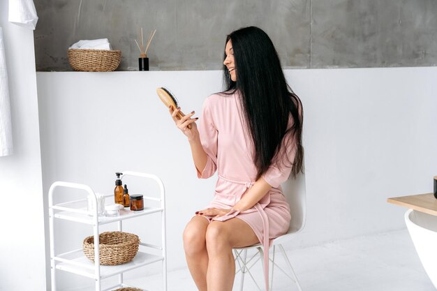 Foto bela caucasiana morena de cabelos longos mulher em um roupão sentar-se em um banheiro no banho segurando um pente de madeira na mão fazendo procedimentos rotineiros de cuidados femininos olhando para a câmera sorrindo