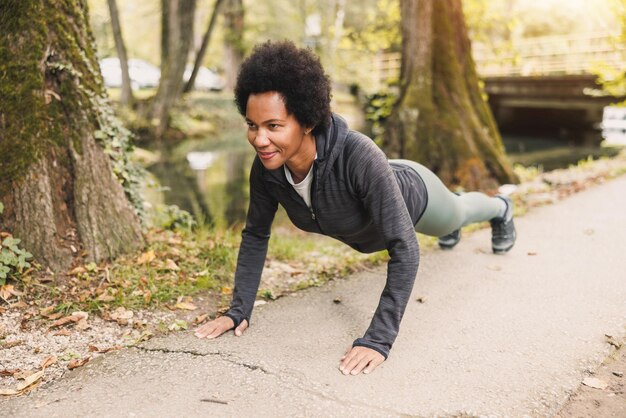 Bela corredora afro-americana madura fazendo exercícios de flexão e se aquecendo para correr na natureza.