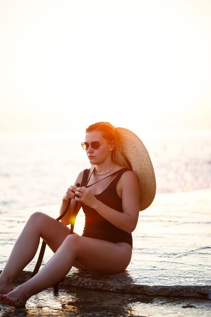 Foto bela dama em um maiô preto à beira-mar, rosto sorridente. luz dourada do pôr do sol