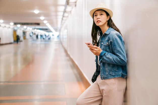 bela dama está esperando namorado na passagem na estação ferroviária. jovem contando com a parede na passagem subterrânea. mulher elegante em pé no corredor usando o aplicativo de comunicação.