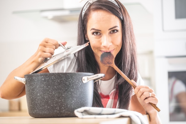 Bela degustação feminina de uma colher de pau, segurando a tampa aberta acima da panela, sorrindo para a câmera.