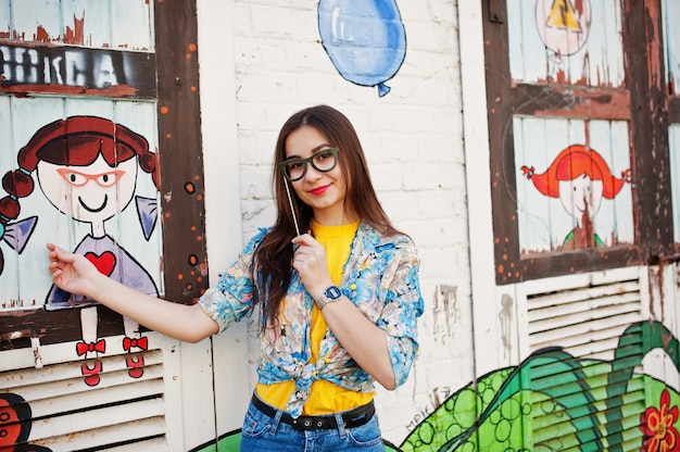 Bela diversão adolescente com óculos na vara usar camiseta amarela, jeans perto de parede grafite.