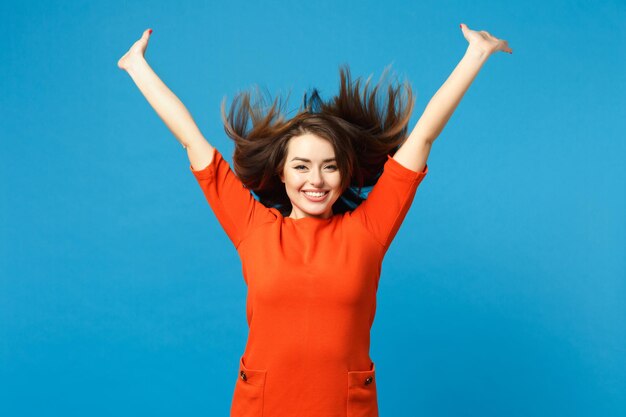 Bela diversão jovem mulher vestindo vermelho vestido laranja brincando esvoaçando cabelo voador levantou as mãos isoladas sobre fundo de parede azul na moda. Conceito de moda de estilo de vida de pessoas. Espaço de cópia de mock up