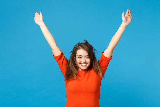 Bela diversão jovem mulher vestindo vermelho vestido laranja brincando esvoaçando cabelo voador levantou as mãos isoladas sobre fundo de parede azul na moda. Conceito de moda de estilo de vida de pessoas. Espaço de cópia de mock up