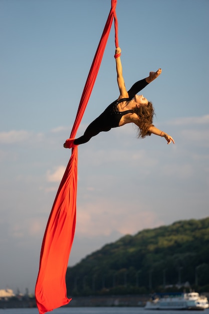 Bela e flexível artista de circo dançando com seda aérea com o céu e a costa do rio