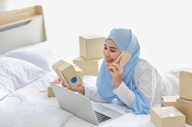 Bela e jovem mulher muçulmana asiática em roupa de noite com aparência atraente, deita-se na cama com computador e entrega de caixa de pacote on-line. mulher freelancer de pequenas empresas de inicialização trabalhando com telefone móvel