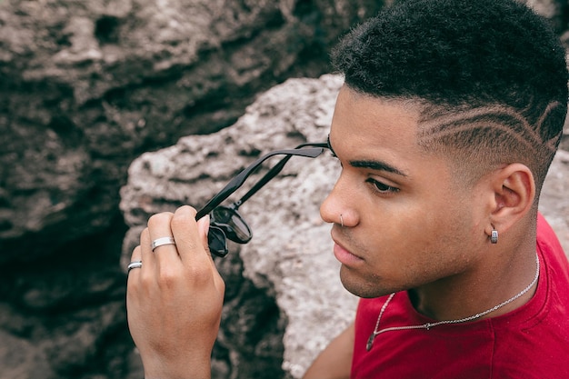 Bela foto de um jovem tirando os óculos escuros enquanto está sentado nas rochas perto do lago