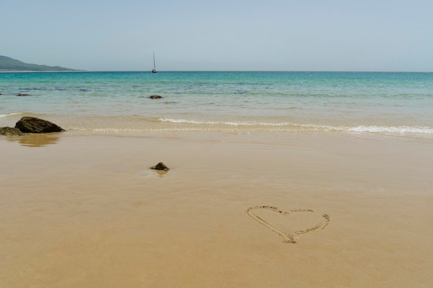 Bela foto de uma praia com um símbolo de coração na areia, águas turquesa e um barco no mar.