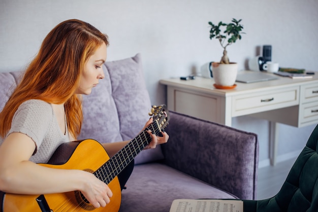 Bela jovem adulta com cabelo comprido, tocando violão, sentado em casa no sofá, foco seletivo. Mulher séria dedilha as cordas, estudando a melodia. Lazer, hobbies, autodesenvolvimento em casa.