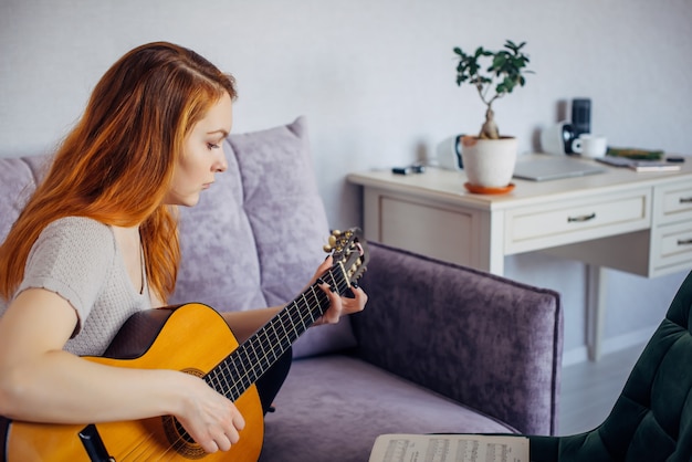 Bela jovem adulta com cabelo comprido, tocando violão, sentado em casa no sofá, foco seletivo. mulher séria dedilha as cordas, estudando a melodia. lazer, hobbies, autodesenvolvimento em casa.