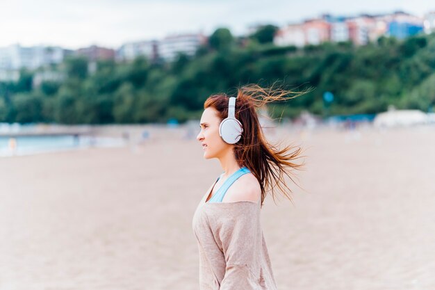 Bela jovem adulta ouvindo música na praia em um dia nublado de verão ou primavera usando fones de ouvido