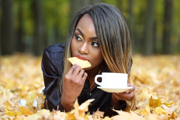 Bela jovem africana bebendo café em uma xícara branca na superfície das folhas de outono no parque