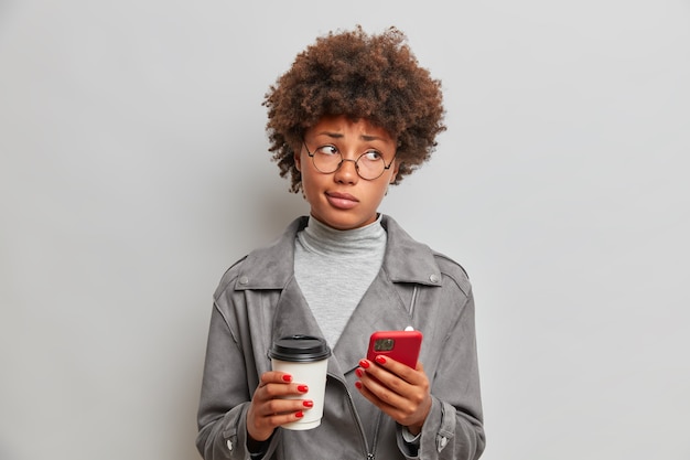 Bela jovem afro-americana, triste e ofendida, parece infeliz, espera por uma chamada importante, usa óculos redondos e jaqueta cinza posa dentro de casa com um gadget moderno