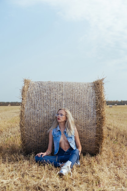 Bela jovem aldeã posando de jeans perto de um fardo de feno em um campo