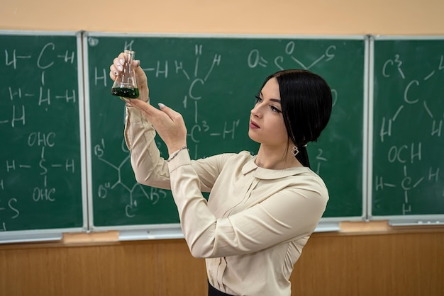 Bela jovem aluna está praticando em uma sala de aula de química com frascos no fundo de uma placa com fórmulas. Conceito de química