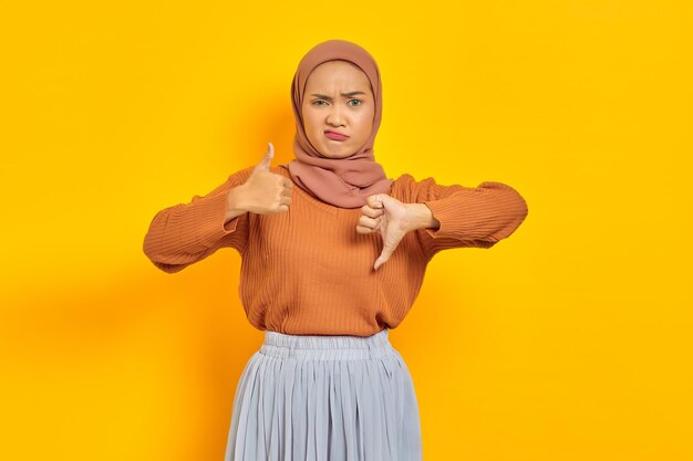 Bela jovem asiática de suéter marrom e hijab mostrando os polegares para cima e para baixo decisão isolada sobre fundo amarelo. Conceito religioso do islamismo de pessoas