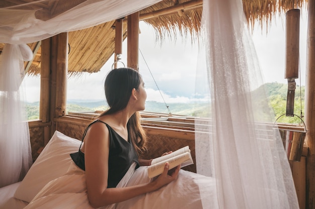 Bela jovem asiática descansando e lendo um livro na cama em uma cabana com teto de palha entre a montanha nas férias