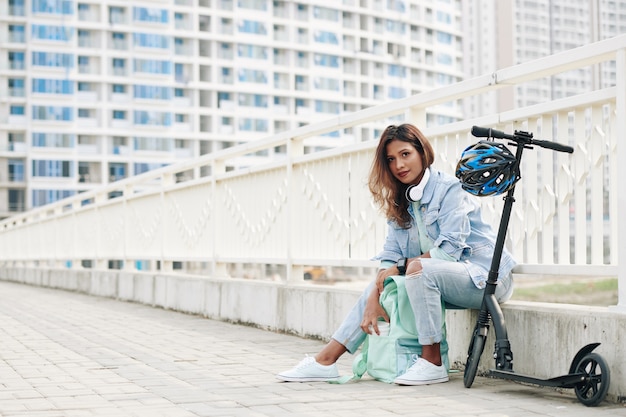 Bela jovem asiática descansando na calçada depois de andar em uma scooter elétrica e tirar uma xícara de café do bolso da mochila