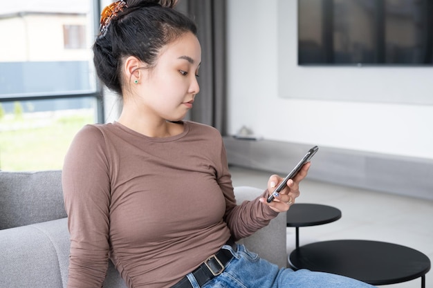 bela jovem asiática em roupas casuais usando telefone sentado no sofá no interior moderno