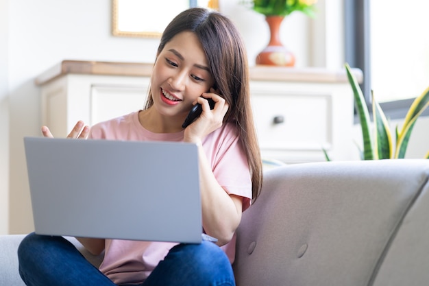 Bela jovem asiática está ligando e usando o computador enquanto está sentada no sofá em casa