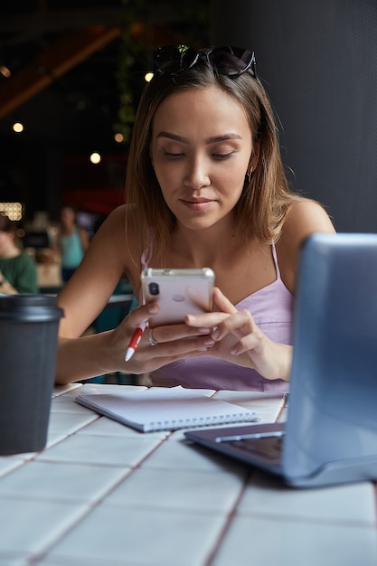 Foto bela jovem asiática sentada com um laptop no café, usando o smartphone