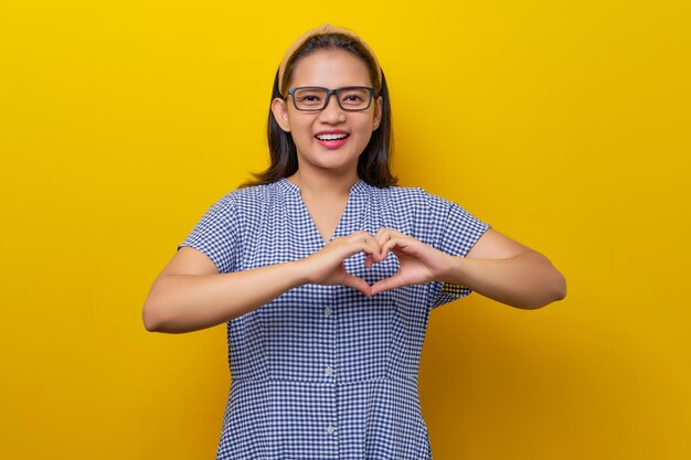 Bela jovem asiática sorridente de 30 anos usando um vestido xadrez com óculos mostrando gesto de coração isolado no conceito de estilo de vida de pessoas de fundo amarelo