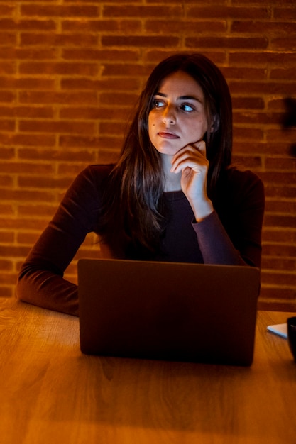 Bela jovem autêntica sentada em uma mesa em uma cozinha aconchegante e usando computador portátil em casa à noite Dúvidas femininas e conversando na Internet e mídias sociais Fazendo pesquisa on-line