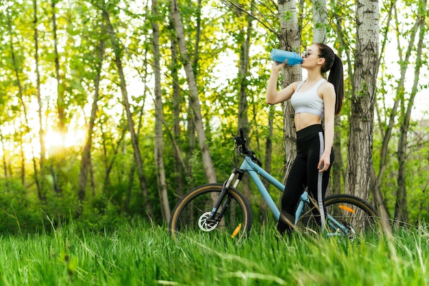 Bela jovem bebe água da coqueteleira enquanto um passeio de bicicleta em um parque no pôr do sol