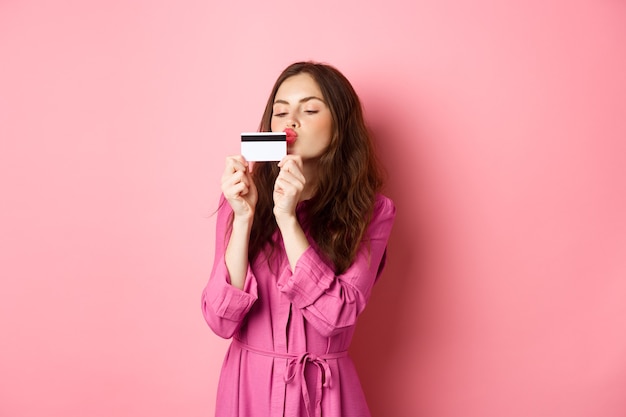 bela jovem beijando o cartão de crédito, indo às compras, desperdiçando dinheiro nas lojas, em pé contra a parede rosa.