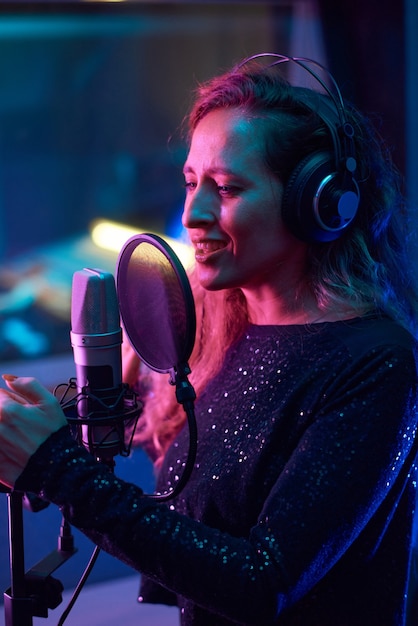 Bela jovem cantora com fones de ouvido cantando uma música para o microfone enquanto está no estúdio de gravação