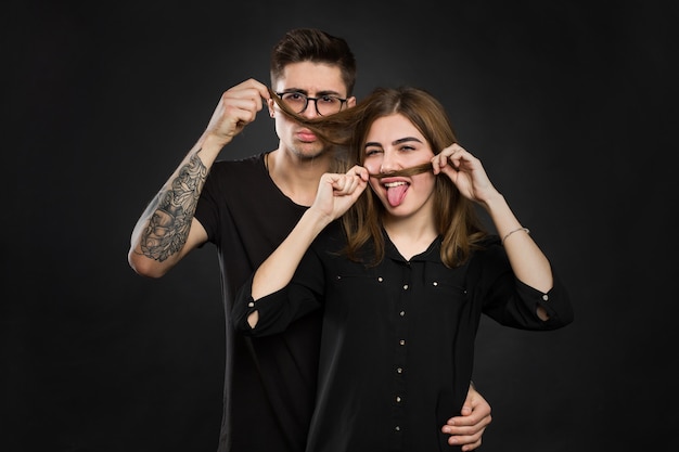 Bela jovem casal apaixonado fazendo bigode falso de cabelo em pé contra um fundo preto. Bigode engraçado.