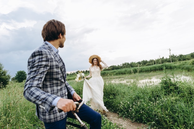 Bela jovem casal noiva e noivo andando em um campo com uma bicicleta