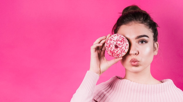 Bela jovem cobrindo os olhos com um delicioso donut cobrindo os olhos dela contra um fundo rosa