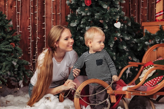 Bela jovem com criança de um ano brincando na árvore de Natal na sala de estar. Mãe com filho bonito na sala decorada de Natal. Eles estão sorrindo e felizes. Conceito de família ano novo. Copie o espaço