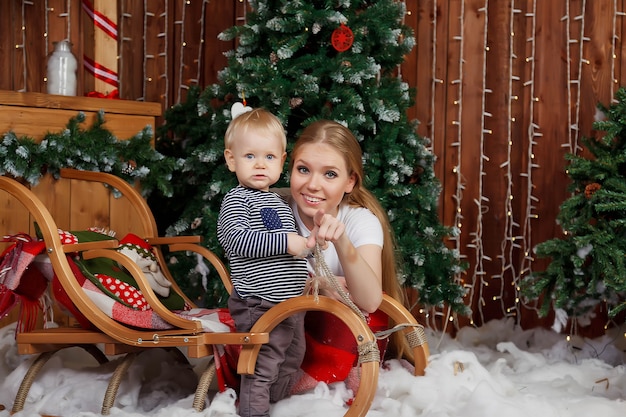 Bela jovem com criança de um ano brincando pela árvore de feliz ano novo. Mãe com filho bonito no quarto decorado de feliz Natal. Eles estão sorrindo e felizes