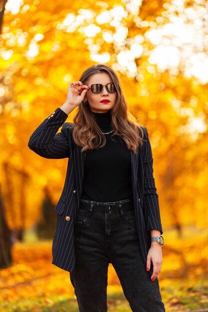 Bela jovem com óculos de sol vintage em roupas da moda, com uma jaqueta elegante, jeans e suéter preto caminha no parque com folhagem de outono laranja ao pôr do sol. elegante estilo feminino de outono