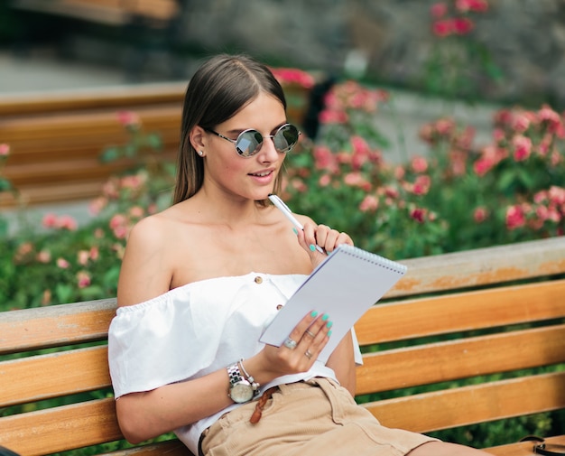Bela jovem com roupas de verão e óculos escuros segurando um smartphone e escrevendo no caderno enquanto está sentado no banco do parque