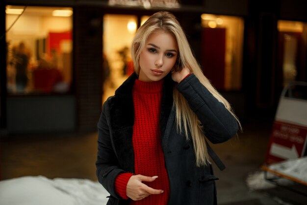 Bela jovem com um casaco elegante e um suéter vermelho posa perto de uma vitrine com luzes